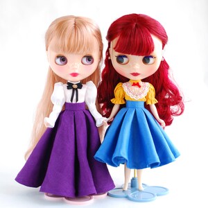 May include: Two dolls wearing different outfits. The doll on the left has blonde hair and is wearing a white blouse with a black bow, a purple skirt, and black shoes. The doll on the right has red hair and is wearing a yellow blouse with a white lace collar, a blue skirt, and white shoes.