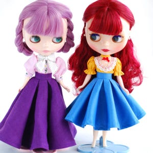 May include: Two dolls wearing dresses. The doll on the left has purple hair and a purple skirt with a pink blouse. The doll on the right has red hair and a blue skirt with a yellow blouse.