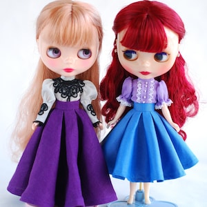 May include: Two dolls wearing dresses. The doll on the left has blonde hair and is wearing a purple dress with a black lace top. The doll on the right has red hair and is wearing a blue dress with a lavender top.