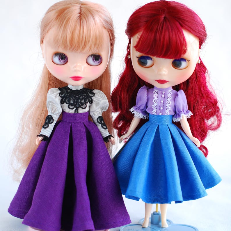 May include: Two dolls wearing dresses. The doll on the left has blonde hair and is wearing a purple dress with a black lace top. The doll on the right has red hair and is wearing a blue dress with a lavender top.
