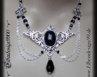 May include: A silver necklace featuring a black gemstone pendant and black beads. The pendant has a filigree design and a teardrop-shaped black crystal hanging from it. The necklace has three chains and is adorned with small black beads.