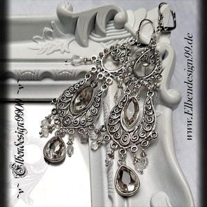 Könnte beinhalten: Silberfarbene Chandelier-Ohrringe mit aufwendigen Filigranarbeiten und klaren Kristallakzenten. Die Ohrringe haben tropfenförmige und oval geschliffene Kristalle, mit kleineren Kristallen, die darunter hängen. Das Design beinhaltet ein dekoratives Rankenmuster.