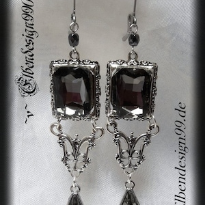 Könnte beinhalten: Ein Paar silberne Ohrringe mit schwarzen tropfenförmigen Kristallen. Die Ohrringe haben einen quadratischen Kristall mit einem silbernen Filigranmuster.