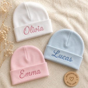 Pode incluir: Três gorros de bebé em branco, azul claro e rosa suave. Cada gorro apresenta uma dobra com nomes bordados: Olivia em rosa, Lucas em azul escuro e Emma em rosa. Uma pequena etiqueta redonda de madeira diz Personalized With Love.