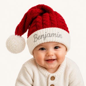 May include: A red and white cable-knit hat with a white pom-pom at the tip. The white ribbed cuff features the name Benjamin embroidered in silver thread. The hat has a classic pointed shape and is made of a soft, textured knit material.
