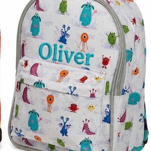 May include: A white backpack featuring a repeating pattern of colourful cartoon monsters in shades of blue, green, orange, red, and purple. The name Oliver is embroidered in turquoise thread on the front pocket, with turquoise embroidery text below.