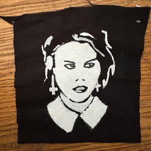 May include: Black and white hand-painted fabric patch featuring a portrait of a person with a white collar and cross earrings.