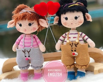 May include: Two crocheted dolls with reindeer designs on their overalls. The doll on the left has pink and grey overalls and brown hair. The doll on the right has brown overalls and black hair. Both dolls are holding red hearts.