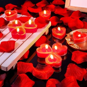 May include: Red heart-shaped candles on a white wooden tray surrounded by red rose petals. The candles are in a variety of sizes and are lit.