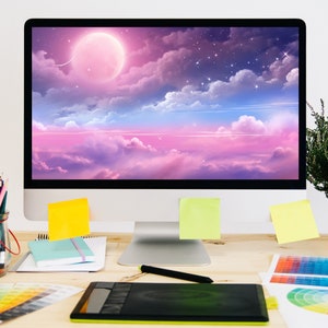 K&ouml;nnte beinhalten: Ein Arbeitsplatz mit einem Computermonitor, der einen rosa und blauen Himmel mit Wolken und einem gro&szlig;en rosa Mond zeigt. Auf dem Schreibtisch befinden sich au&szlig;erdem Farbmuster, ein Grafiktablett, Bleistifte und eine Pflanze.