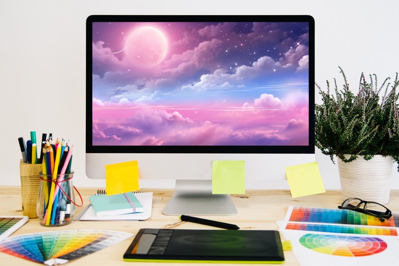 K&ouml;nnte beinhalten: Ein Arbeitsplatz mit einem Computermonitor, der einen rosa und blauen Himmel mit Wolken und einem gro&szlig;en rosa Mond zeigt. Auf dem Schreibtisch befinden sich au&szlig;erdem Farbmuster, ein Grafiktablett, Bleistifte und eine Pflanze.