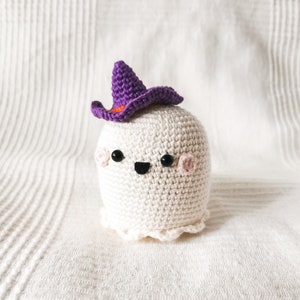 May include: A white crocheted ghost wearing a purple witch hat with an orange brim. The ghost has black eyes and a black smiling mouth.