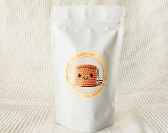 May include: A white pouch with a circular sticker featuring a crocheted basket with a smiling face. The sticker reads "Baby Shumoi - DIY Crochet Kit" and "Emi Creations by Chloe".