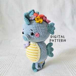 May include: A crocheted seahorse amigurumi toy with a blue body, yellow and white striped belly, and a colourful flower crown. The toy is standing on a white surface. Digital Pattern.