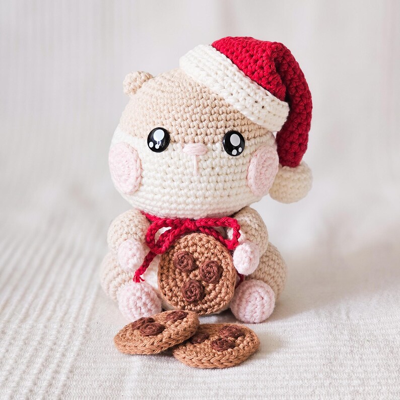 Puede incluir: Un h&aacute;mster de ganchillo con un gorro de Pap&aacute; Noel rojo y blanco y sosteniendo una galleta de ganchillo. El h&aacute;mster es beige con mejillas rosadas y grandes ojos negros. Dos galletas de ganchillo adicionales est&aacute;n delante.