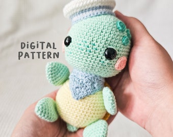 May include: A crocheted turtle amigurumi toy wearing a blue and white sailor hat. The turtle is green, yellow, and blue. The toy is being held in a hand.