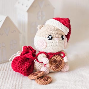Puede incluir: Un h&aacute;mster de ganchillo con gorro y bufanda de Pap&aacute; Noel, sosteniendo una galleta y una bolsa roja con cord&oacute;n. Dos galletas adicionales est&aacute;n frente al h&aacute;mster. El h&aacute;mster es beige, blanco y rosa, con ojos negros. El gorro y la bolsa son rojos.