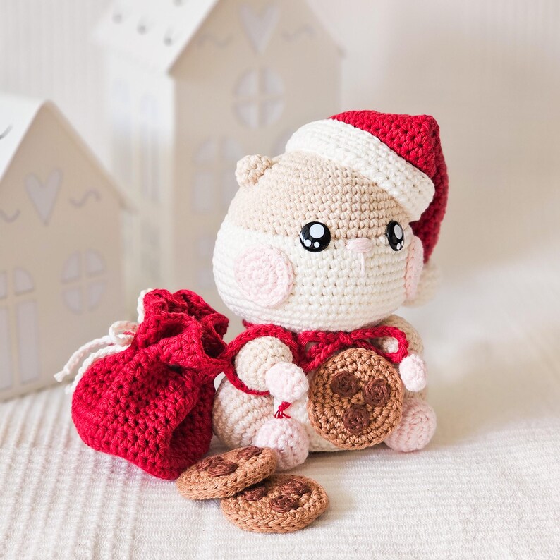 Puede incluir: Un h&aacute;mster de ganchillo con gorro y bufanda de Pap&aacute; Noel, sosteniendo una galleta y una bolsa roja con cord&oacute;n. Dos galletas adicionales est&aacute;n frente al h&aacute;mster. El h&aacute;mster es beige, blanco y rosa, con ojos negros. El gorro y la bolsa son rojos.