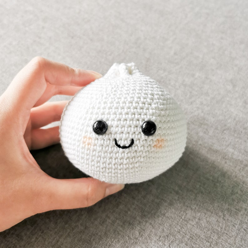 May include: A white crocheted dumpling with a smiling face, black eyes, and pink cheeks.