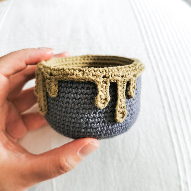 May include: A small crocheted bowl with a gray base and a brown rim. The rim is crocheted in a dripping pattern.