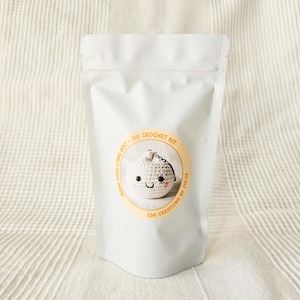 May include: A white pouch with a circular sticker featuring a crocheted dumpling with a smiling face. The sticker reads "Baby Xiao Long Bao - DIY Crochet Kit" and "Emi Creations by Chloe."
