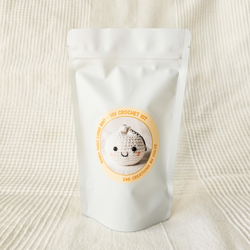 May include: A white pouch with a circular sticker featuring a crocheted dumpling with a smiling face. The sticker reads "Baby Xiao Long Bao - DIY Crochet Kit" and "Emi Creations by Chloe."