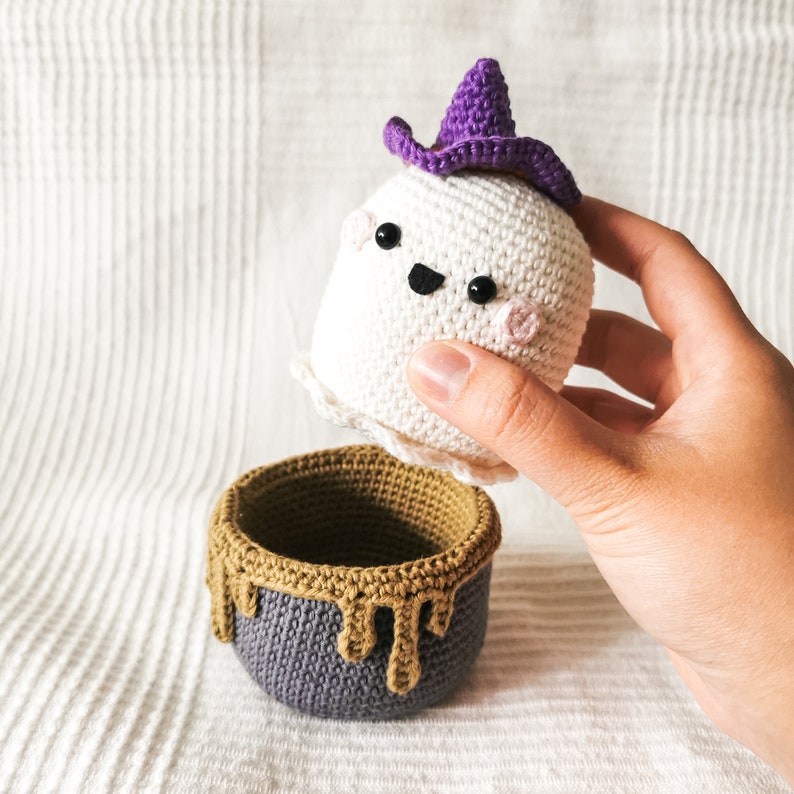 May include: A crocheted ghost with a purple witch hat, holding a crocheted cauldron with a brown rim and gray body.
