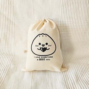 May include: A beige drawstring bag with a black illustration of a dumpling with a heart and the text "I LOVE EVERYTHING A-BAO YOU". The bag is made of a soft, natural fabric.