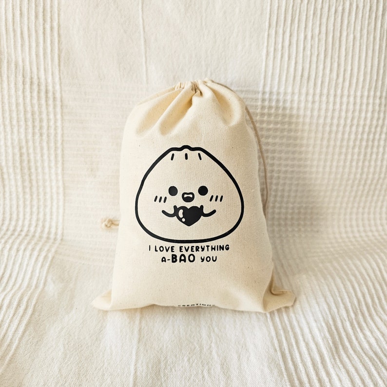 May include: A beige drawstring bag with a black illustration of a dumpling with a heart and the text "I LOVE EVERYTHING A-BAO YOU". The bag is made of a soft, natural fabric.
