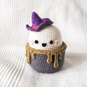 May include: A crocheted ghost wearing a purple witch hat with an orange brim. The ghost is sitting in a gray cauldron with a brown crocheted rim that looks like dripping chocolate.