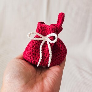 Puede incluir: Una peque&ntilde;a bolsa roja tejida a mano con un cierre de cord&oacute;n blanco. La bolsa se sostiene en una mano, mostrando la textura del hilo y el delicado cord&oacute;n blanco. La bolsa mide aproximadamente 5 cm de alto.