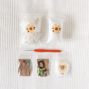 May include: A crochet kit with various supplies. The kit includes white yarn, stuffing, an orange crochet hook, butterfly pins, and a practice bag. The items are packaged in clear plastic bags and arranged on a white surface.