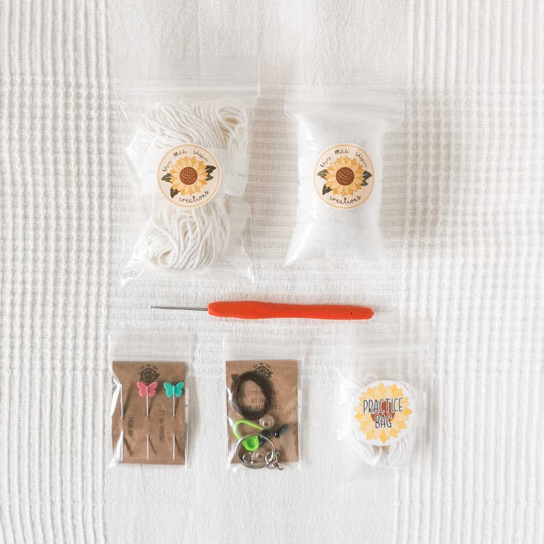 May include: A crochet kit with various supplies. The kit includes white yarn, stuffing, an orange crochet hook, butterfly pins, and a practice bag. The items are packaged in clear plastic bags and arranged on a white surface.