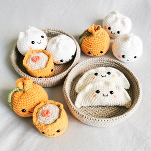 Könnte beinhalten: Handgemachtes Häkel-Essensset mit lächelnden Knödeln, Brötchen und Sushi in beigen gewebten Körben. Das Set umfasst weiße und orangefarbene Lebensmittel mit gestickten Details, ideal für eine Kinderküche oder als dekorative Artikel.