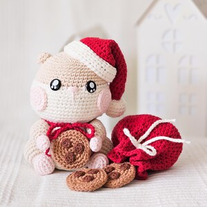 Puede incluir: Un h&aacute;mster de ganchillo con un gorro de Pap&aacute; Noel rojo y blanco y una galleta con chispas de chocolate. Una bolsa roja con cord&oacute;n y dos galletas m&aacute;s est&aacute;n cerca. El h&aacute;mster tiene mejillas rosas y un lazo rojo.