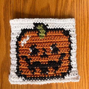 May include: A square crochet coaster featuring a jack-o'-lantern design. The pumpkin is orange with a black stitched face and a green stem, all set against a white border. The texture is created with visible yarn loops in a grid pattern.