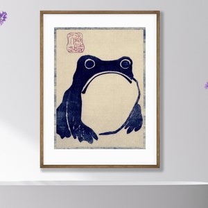 Puede incluir: Una impresión enmarcada de una rana azul en estilo xilografía japonés. La rana está sentada con la boca cerrada y los ojos cerrados. La impresión está sobre un fondo de color crema con un sello rojo en la esquina superior izquierda.