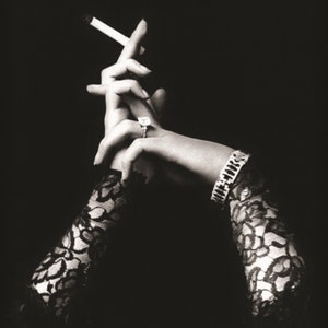 Pode incluir: Um par de mãos com luvas de renda preta, uma segurando um cigarro, a outra com um anel no dedo. As mãos estão postas sobre um fundo escuro.