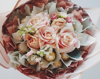 May include: A bouquet of pink roses and white flowers with a center of chocolate candies wrapped in gold foil.