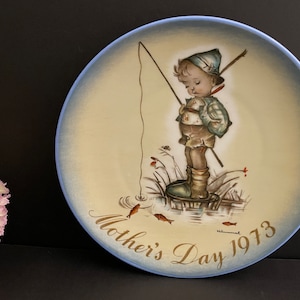 Può includere: Un piatto in ceramica blu e bianco con un dipinto di un ragazzo che pesca. Il piatto ha il testo "Mother's Day 1973" sul fondo.