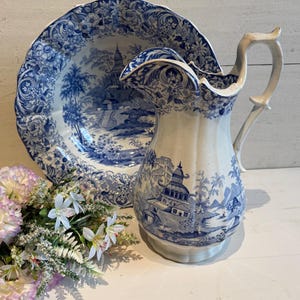 Può includere: Una brocca in ceramica bianca e una grande bacinella abbinata, entrambe decorate con un motivo blu a trasferimento raffigurante pagode, alberi e bordi floreali. La brocca ha un manico curvo e un bordo smerlato, la bacinella è scanalata.
