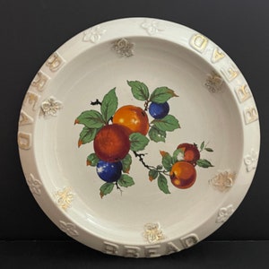 May include: White ceramic bread plate with a gold rim and the word "BREAD" embossed around the edge. The centre of the plate features a painted design of a branch with red, orange, and blue fruit.