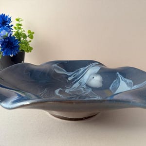 May include: A dark blue ceramic bowl with a wavy rim and a white and blue marbled design. The bowl is on a light beige surface. A small black vase with blue flowers is in the background.