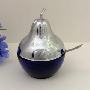 Può includere: Una zuccheriera a forma di pera con coperchio argentato lucido e base blu scuro. Un cucchiaino d'argento poggia sul bordo. Un oggetto decorativo per la cucina.