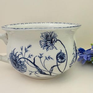 May include: White ceramic bowl with a handle, adorned with blue floral patterns. The bowl has a wide opening and a slightly distressed rim. A sprig of blue flowers sits to the right of the bowl. This decorative bowl is ideal for home decor.