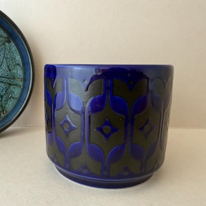 May include: A cobalt blue ceramic planter with a black geometric pattern. The planter has a cylindrical shape and a slightly flared base. A blue and green decorative plate is partially visible in the background.