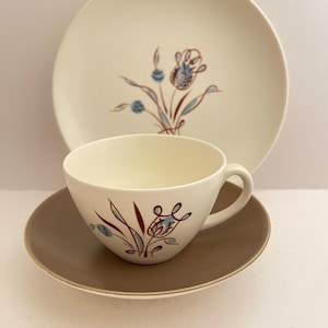 Può includere: Servizio da tè vintage color crema. La tazza, il piattino e il piatto hanno un bordo marrone e un motivo floreale blu e marrone. Il piatto è dietro la tazza e il piattino.