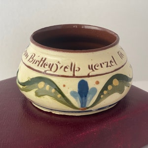 Puede incluir: Un pequeño cuenco redondo de cerámica con exterior color crema e interior marrón. El cuenco está decorado con motivos florales verdes y azules y tiene texto alrededor del borde. El cuenco está sobre una superficie burdeos.