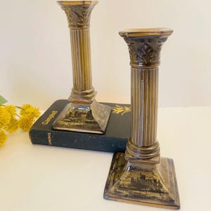 May include: A pair of gold ceramic candle holders shaped like columns with a detailed design. The bases of the holders feature a black and white image of a ship.