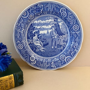 May include: A round ceramic plate featuring a blue and white transferware pattern. The central scene depicts a person sitting with a child and another person standing with a tool, surrounded by a border of floral motifs and geometric patterns.